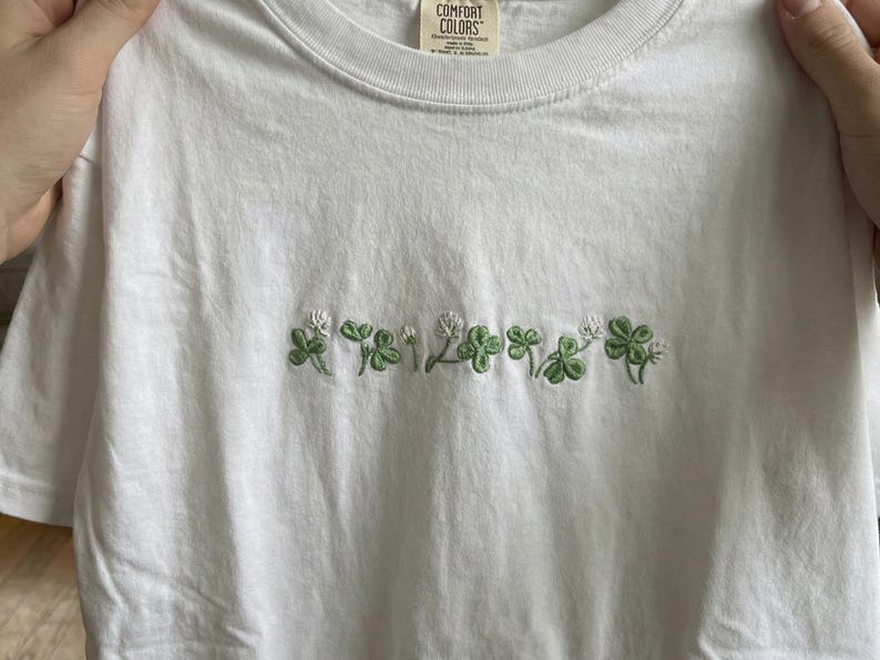 Pu&ograve; includere: T-shirt bianco con trifogli verdi ricamati e fiori bianchi sul petto. La maglietta &egrave; realizzata in un materiale morbido e confortevole. L'etichetta recita "Comfort Colors".