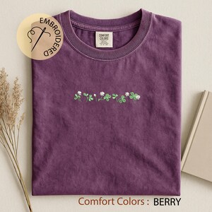 Pu&ograve; includere: Una maglietta color bacca con scollo tondo, con un ricamo floreale di fiori bianchi e foglie verdi. La maglietta ha un'etichetta Comfort Colors e la parola "EMBROIDERED" in un cerchio.