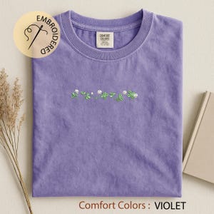 Pu&ograve; includere: Una maglietta viola con scollo tondo, con un ricamo di trifogli verdi e fiori bianchi. La maglietta ha un'etichetta con la scritta "Comfort Colors". Il testo "Comfort Colors: VIOLET" &egrave; in basso.