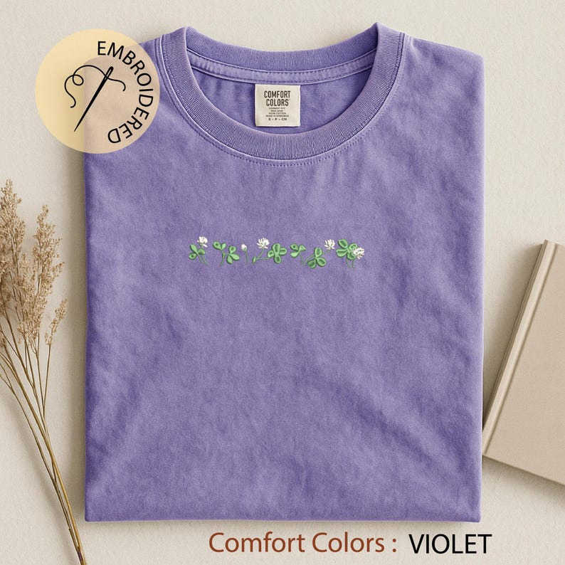 Pu&ograve; includere: Una maglietta viola con scollo tondo, con un ricamo di trifogli verdi e fiori bianchi. La maglietta ha un'etichetta con la scritta "Comfort Colors". Il testo "Comfort Colors: VIOLET" &egrave; in basso.