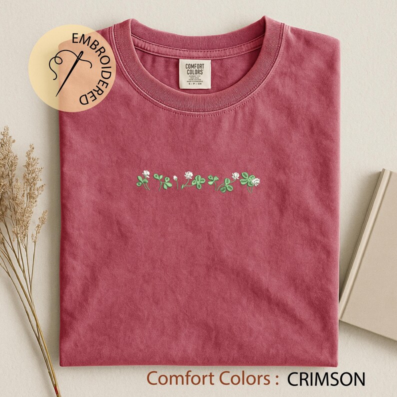 Pu&ograve; includere: T-shirt color cremisi con ricami di trifogli verdi e fiori bianchi. La maglietta ha uno scollo tondo e una piccola etichetta sul colletto. Il testo "Comfort Colors : CRIMSON" &egrave; nella parte inferiore dell'immagine.