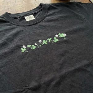 Pu&ograve; includere: T-shirt grigio scuro con ricamo di trifogli. I trifogli sono verdi con fiori bianchi. La maglietta ha uno scollo tondo e un'etichetta con la scritta "Comfort Colors".