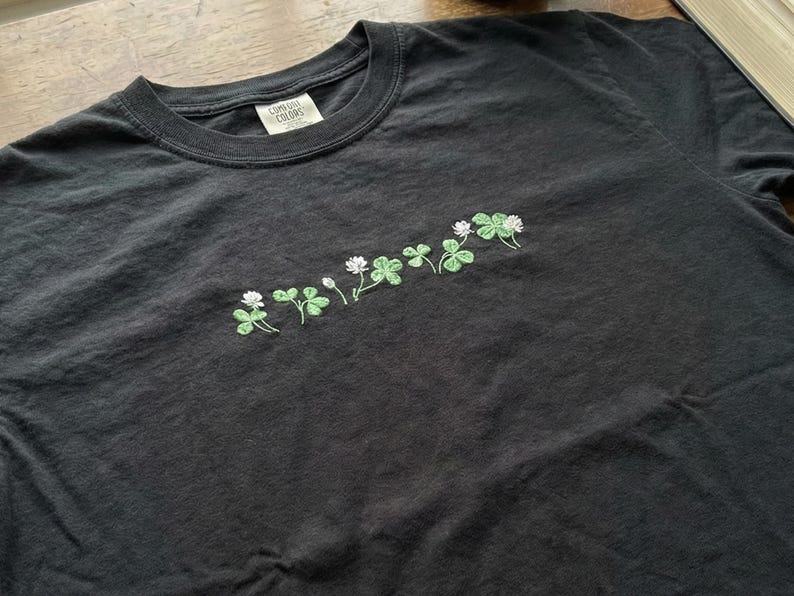 Pu&ograve; includere: T-shirt grigio scuro con ricamo di trifogli. I trifogli sono verdi con fiori bianchi. La maglietta ha uno scollo tondo e un'etichetta con la scritta "Comfort Colors".