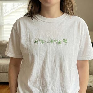 Pu&ograve; includere: T-shirt bianca con trifogli verdi ricamati e la scritta "124". La maglietta a maniche corte &egrave; realizzata in un materiale morbido e confortevole. Il disegno &egrave; centrato sul petto.