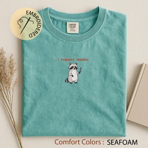K&ouml;nnte beinhalten: T&uuml;rkisgr&uuml;nes T-Shirt mit dem gestickten Schriftzug "I require snacks" &uuml;ber einem Cartoon-Waschb&auml;ren, der ein Messer h&auml;lt. Das Shirt ist mit "Comfort Colors" gekennzeichnet und hat eine einfarbige Farbe.