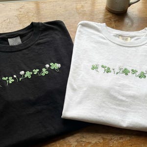 Pu&ograve; includere: Due magliette, una nera e una bianca, ciascuna con girocollo e design a trifogli verdi e bianchi ricamati. Le magliette sono realizzate in un materiale morbido e probabilmente per un abbigliamento casual.