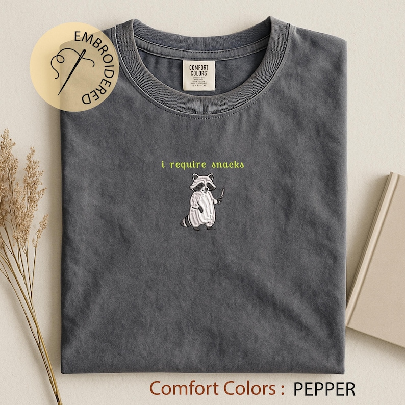 K&ouml;nnte beinhalten: Ein graues Comfort Colors T-Shirt mit dem gestickten Text "I require snacks" und einer Waschb&auml;r-Grafik, die ein Messer h&auml;lt. Das Shirt hat einen Rundhalsausschnitt und kurze &Auml;rmel. Das Shirt ist pfefferfarben.