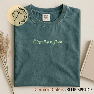 Pu&ograve; includere: Una maglietta Comfort Colors blu abete con ricami di trifogli bianchi e verdi. La maglietta ha un girocollo e una piccola etichetta sul colletto. Il testo "Comfort Colors : BLUE SPRUCE" &egrave; in basso nell'immagine.