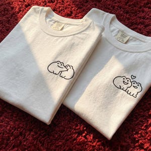 May include: Two white t-shirts with black frog graphics. One shirt features two frogs hugging, the other shows two frogs with a heart above them. The shirts are folded and rest on a red surface.