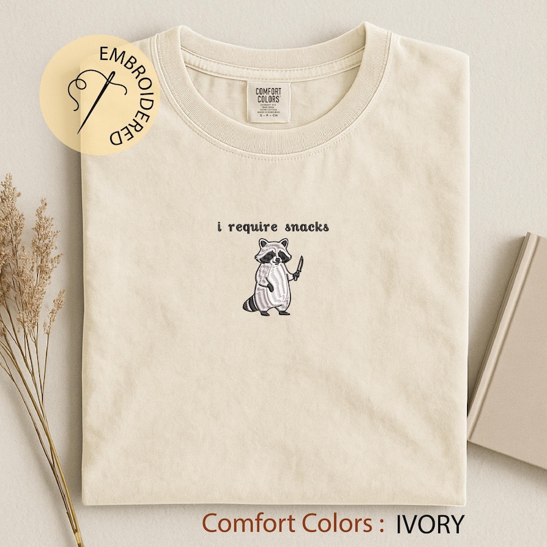 K&ouml;nnte beinhalten: Elfenbeinfarbenes Comfort Colors T-Shirt mit dem gestickten Text "i require snacks" &uuml;ber einem Cartoon-Waschb&auml;ren, der ein Messer h&auml;lt. Das Shirt hat einen Rundhalsausschnitt und ein kleines Etikett. Die Worte "Comfort Colors : IVORY" stehen unten.