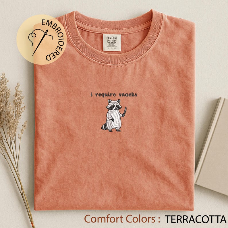 K&ouml;nnte beinhalten: Terracottafarbenes T-Shirt mit der gestickten Aufschrift "i require snacks" und einem Waschb&auml;ren mit Messer. Das Shirt ist von der Marke Comfort Colors und hat einen Rundhalsausschnitt.