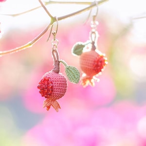 May include: A pair of dangling earrings featuring miniature crocheted pomegranate fruits. Each pomegranate is crafted with textured pink and red thread, accented by a small green crocheted leaf and a brown stem, suspended from silver-toned metal hooks.