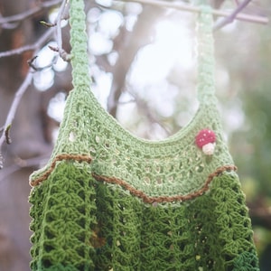 May include: A handmade green crochet top with a brown trim and a small mushroom embellishment. The top is hanging from a tree branch, showcasing its intricate stitchwork and earthy tones. The crochet design features a textured pattern and a scoop neckline.