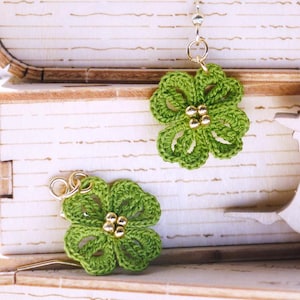May include: A pair of green, four-leaf clover earrings with gold-colored beads and hooks. The earrings are displayed on a stack of wooden book-shaped boxes. A decorative wooden cutout is also visible.