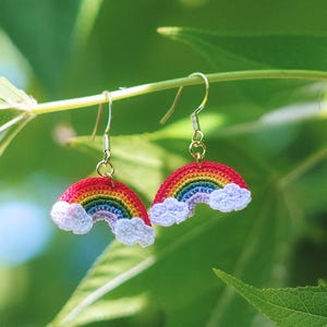 May include: A pair of rainbow-shaped earrings with red, orange, yellow, green, blue, and purple arches, and white cloud accents. The earrings are suspended from gold-colored hooks, set against a backdrop of green leaves.