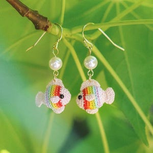 May include: A pair of dangle earrings featuring small, crocheted fish charms with rainbow-colored stripes, white fins, and black eyes. Each fish is suspended from a gold-toned hook by a single white pearl bead, creating a delicate and colorful accessory.