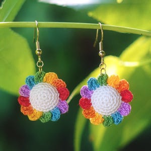 May include: A pair of handmade crochet flower earrings. Each earring features a white center surrounded by colorful petals in shades of red, orange, yellow, green, blue, and purple. The earrings hang from gold-colored hooks.