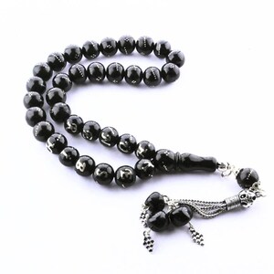 Peut inclure: Chapelet noir avec des détails argentés. Les perles sont rondes et lisses, reliées par un pompon. Les perles ont des marquages argentés. Le pompon est argenté avec un motif décoratif.