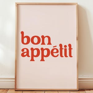 Może przedstawiać: Oprawiony wydruk artystyczny z napisem "bon appétit" w pogrubionej, czerwonej czcionce na bladym różowym tle. Drewniana rama uzupełnia ciepłe odcienie wydruku, który opiera się o białą ścianę i drewnianą podłogę.