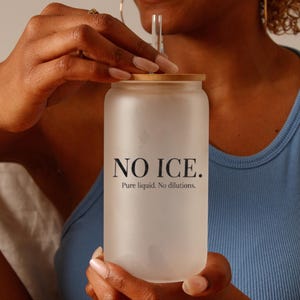 May include: A frosted glass tumbler with a bamboo lid and a clear straw. The tumbler has the text "NO ICE. Pure liquid. No dilutions." printed in black. The person is wearing a blue vest top.