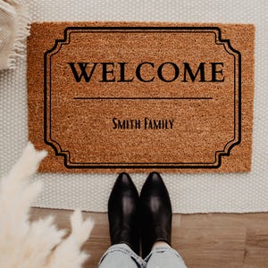 May include: A rectangular doormat made of brown coir fiber, featuring the word "WELCOME" in black, above the text "SMITH FAMILY". The mat has a decorative black border and is placed on a white rug. Black boots and blue jeans are visible.