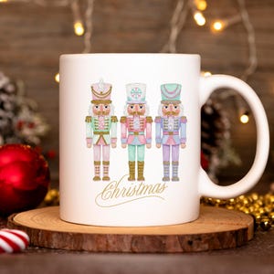 May include: White ceramic mug with three pastel-coloured nutcracker illustrations in pink, mint, and lavender. The word "Christmas" is written in gold script below the nutcrackers. The mug has a curved handle and is set on a wooden coaster.