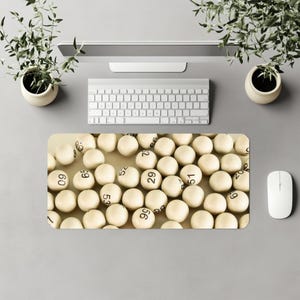 May include: A computer desk mat featuring a beige background filled with white bingo balls, each with a black number. The mat is surrounded by a keyboard, mouse, and two small potted plants, all on a gray surface.