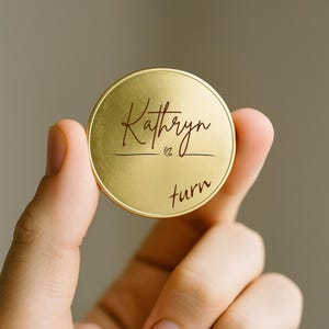 May include: A gold-colored round token with the name "Kathryn" in a cursive font, with the word "turn" below. The token is held between fingers, with a blurred background.