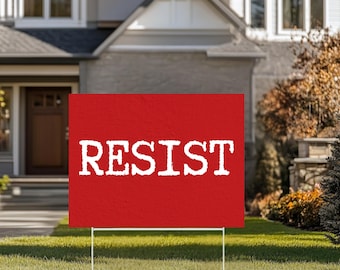May include: A red garden sign displaying the word "RESIST" in white, set on a lawn in front of a house. The sign is supported by a white metal stake.