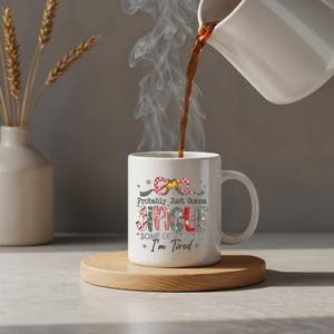 May include: A white ceramic mug with the text "Probably Just Gonna Jingle Some of the Way * I'm Tired" in a festive design. The mug is on a wooden coaster, with coffee being poured into it. A vase with dried wheat is in the background.