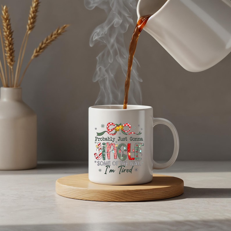 May include: A white ceramic mug with the text "Probably Just Gonna Jingle Some of the Way * I'm Tired" in a festive design. The mug is on a wooden coaster, with coffee being poured into it. A vase with dried wheat is in the background.