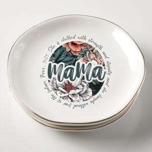 May include: Three white ceramic plates with a gold rim, stacked together. Each plate has a floral design with the word "MAMA" in the centre. The plates have a wavy shape and are suitable for serving food.
