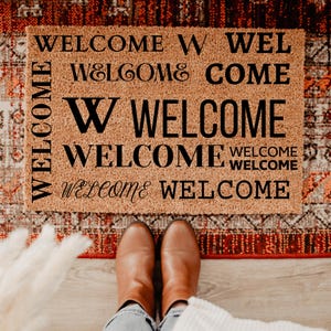 May include: A rectangular doormat with the word "WELCOME" in various fonts and arrangements. The mat is a natural brown color, with black text. The mat is on a patterned rug, and a person is wearing brown boots.