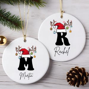 May include: Two white ceramic Christmas ornaments. Each ornament features a black letter, a red Santa hat with antlers, and a name. One ornament has the letter "M" and the name "Martin". The other has the letter "R" and the name "Rachel".