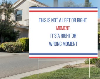 May include: A white sign with red, white, and blue stripes at the top and bottom, displayed on a lawn. The sign reads, "THIS IS NOT A LEFT OR RIGHT MOMENT, IT'S A RIGHT OR WRONG MOMENT" in blue text. A house and blue sky are in the background.