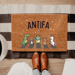 May include: A brown coir doormat with the word "ANTIFA" in navy blue. The mat features a cartoon illustration of a dinosaur, raccoon, frog, bear, and unicorn crossing a crosswalk. The mat is placed on a black and white striped rug.