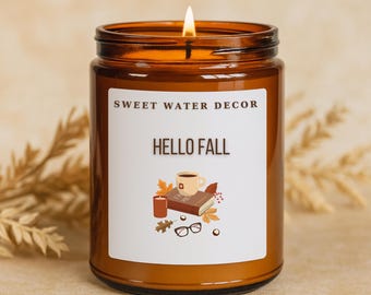 May include: A lit brown glass jar candle with a white label that reads "SWEET WATER DECOR" and "HELLO FALL". The label features a graphic of a coffee cup, book, candle, glasses, and fall leaves. The candle is surrounded by dried wheat.