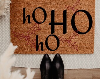 May include: A rectangular coir doormat with the words "ho ho ho" in black, and red holly accents. The mat is brown. Black boots and blue jeans are visible at the bottom of the image. A festive welcome mat.