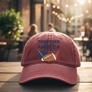 May include: A maroon baseball cap featuring the text "TOGETHER WE ARE AMERICA" above an American flag and a football. The cap is made of a soft material and has a curved brim. The background is blurred, suggesting an outdoor setting.