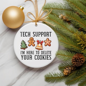 May include: A white ceramic Christmas ornament with the text "TECH SUPPORT" and "I'M HERE TO DELETE YOUR COOKIES". The ornament features gingerbread-themed graphics. A gold ornament and pine branches are in the background.
