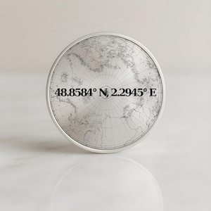 Peut inclure: Un bouton rond argenté avec une carte du monde détaillée gravée en gris et blanc. Les coordonnées 48.8584° N, 2.2945° E sont imprimées en noir. Le bouton a une surface réfléchissante.