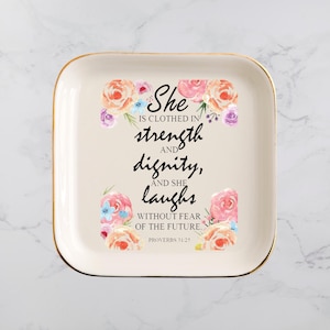 May include: A square, cream-coloured ceramic tray with a gold rim. The tray features floral accents and the text "She is clothed in strength and dignity, and she laughs without fear of the future." Proverbs 31:25.