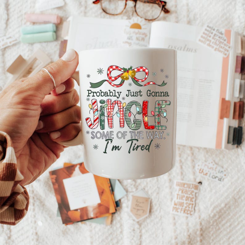 May include: White ceramic mug with the text "Probably Just Gonna Jingle Some of the Way. I'm Tired" in a festive font. The mug features a red and green plaid bow and Christmas lights design, perfect for the holiday season.