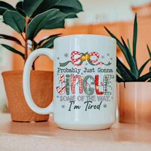 May include: White ceramic mug with a large handle, featuring the text "Probably Just Gonna Jingle Some Of The Way. I'm Tired" in a festive font. The design includes a red bow, bells, and Christmas lights.