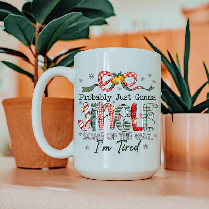 May include: White ceramic mug with a large handle, featuring the text "Probably Just Gonna Jingle Some Of The Way. I'm Tired" in a festive font. The design includes a red bow, bells, and Christmas lights.