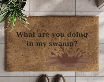 May include: A rectangular doormat made of natural brown fibers. The mat features the phrase "What are you doing in my swamp?" in dark green text, along with a brown mud splash graphic. Brown leather shoes are at the bottom.