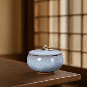 May include: A small, round, light blue ceramic jar with a lid and a brass ring handle. The jar has a textured, crackled surface and a reddish-brown base. The lid is slightly domed and the brass ring is attached to the top.