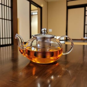 Könnte beinhalten: Eine klare Glas-Teekanne mit einem Deckel und einem Infuser aus Edelstahl, gefüllt mit bernsteinfarbenem Tee. Die Teekanne hat einen runden Körper und einen gebogenen Griff und Ausguss. Der Tee ist durch das Glas sichtbar, und die Teekanne steht auf einer dunklen Holzoberfläche.