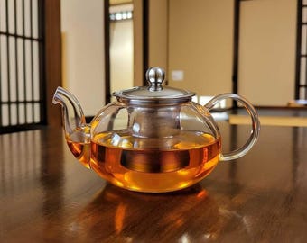 May include: A clear glass teapot with a stainless steel lid and infuser, filled with amber-colored tea. The teapot has a rounded body and a curved handle and spout. The tea is visible through the glass, and the teapot sits on a dark wooden surface.