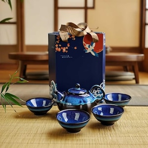 Puede incluir: Un juego de té de cerámica azul con una tetera y cuatro tazas a juego. La tetera y las tazas tienen un acabado brillante con una combinación de colores azul oscuro y azul claro. Una caja de regalo con una ilustración de grulla está detrás del juego de té.